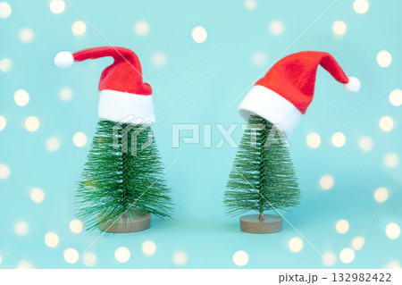 Two small Christmas trees in red Santa Hats. Decorating of Artificial Fir tree. Beautiful imitation of a mini Christmas tree, with Santa Cap on blue background. Christmas holidays concept. Minimalism. 132982422