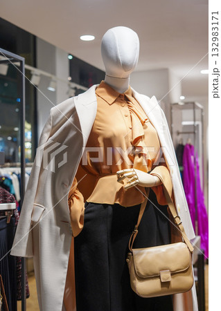 Elegant female mannequin dressed in beige blouse, white coat, and handbag in store Elegant female mannequin dressed in beige blouse, white coat, and handbag in store 132983171
