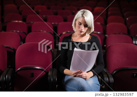 Woman wearing fitted black top reading script pages while sitting in red theater seats Woman wearing fitted black top reading script pages while sitting in red theater seats 132983597