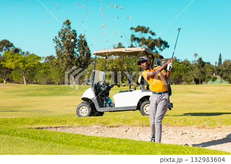 African American male golfer swinging club from sand bunker sending ball in orange polo, copy space African American male golfer swinging club from sand bunker sending ball in orange polo, copy space 132983604