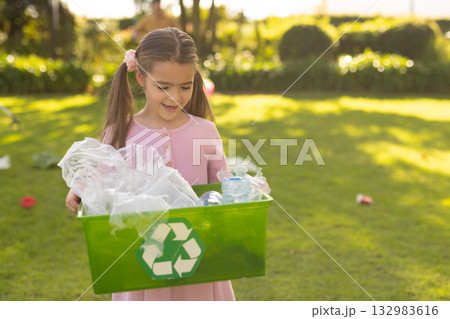 Girl wearing pink dress standing on grass holding green recycling bin with plastic bottles and bags 132983616