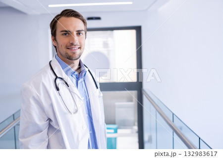 Male doctor standing in bright hospital corridor holding stethoscope near glass railing 132983617