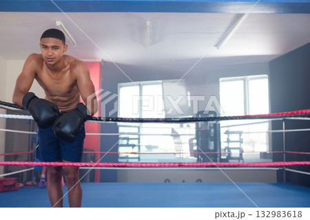 African American male boxer in gym wearing gloves leaning on multicolored ropes on blue ring mat African American male boxer in gym wearing gloves leaning on multicolored ropes on blue ring mat 132983618