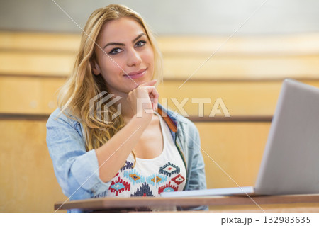 Female student sitting at lecture hall desk resting chin on hand studying with papers and laptop Female student sitting at lecture hall desk resting chin on hand studying with papers and laptop 132983635