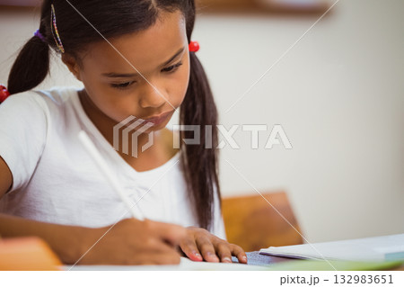 Girl student holding pencil and writing in open notebook at wooden desk in classroom Girl student holding pencil and writing in open notebook at wooden desk in classroom 132983651