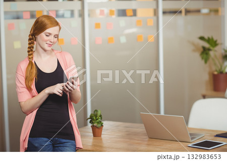 Red-haired woman standing by table in office checking phone near succulent and notes, copy space Red-haired woman standing by table in office checking phone near succulent and notes, copy space 132983653