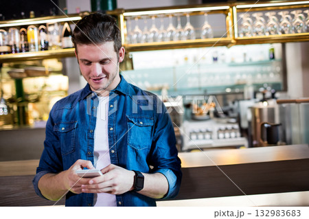 Man checking smartphone at cafe counter wearing smartwatch with glassware and bottles on shelves 132983683