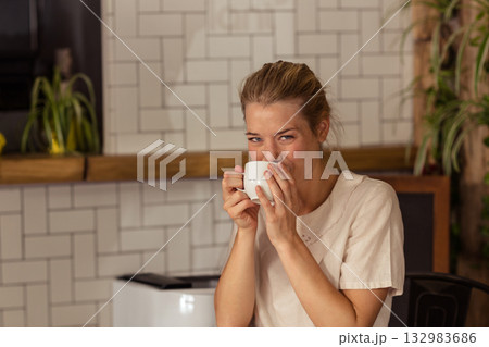 Woman sipping from white ceramic mug near mouth at cafe corner with coffee machine, potted plants Woman sipping from white ceramic mug near mouth at cafe corner with coffee machine, potted plants 132983686