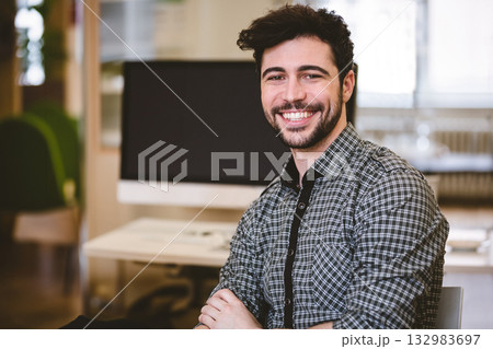 Man sitting at desk crossing arms in office workspace wearing checkered shirt with computer monitor Man sitting at desk crossing arms in office workspace wearing checkered shirt with computer monitor 132983697