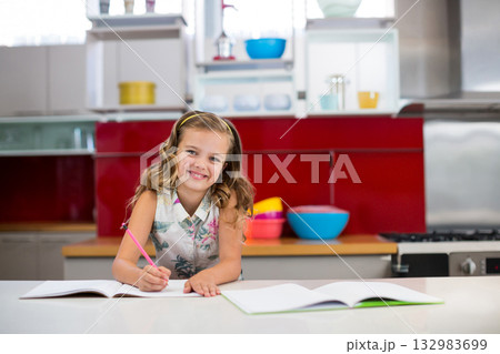 Girl writing in open notebook on kitchen countertop using pink pencil near bowls and refrigerator Girl writing in open notebook on kitchen countertop using pink pencil near bowls and refrigerator 132983699