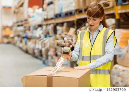 Female worker in hi-vis vest sealing cardboard box with tape dispenser in storage aisle, copy space 132983709