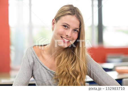 Smiling teenage girl sitting at classroom desk near chairs under large windows with red walls 132983710