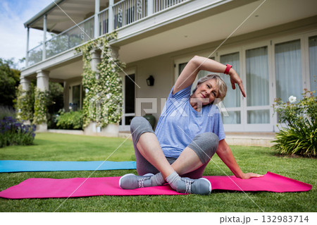 Senior woman stretching on pink, blue yoga mats in front yard with fitness tracker, copy space 132983714
