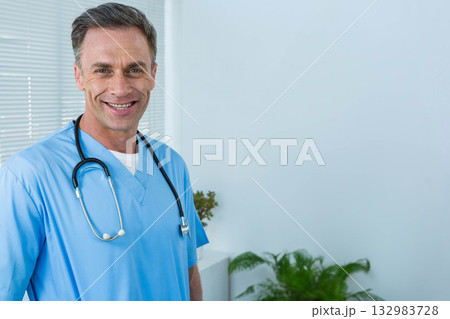 Male doctor standing and smiling at viewer in clinic with blue scrubs, stethoscope, copy space 132983728
