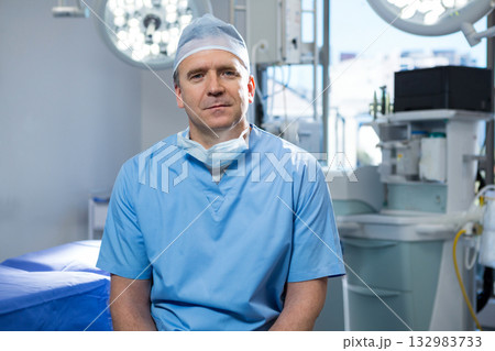 Male surgeon sitting in surgery room in scrubs beside anesthesia machine under overhead lights Male surgeon sitting in surgery room in scrubs beside anesthesia machine under overhead lights 132983733