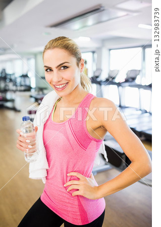 Woman in twenties smiling in gym wearing sportswear holding water bottle with towel by treadmills Woman in twenties smiling in gym wearing sportswear holding water bottle with towel by treadmills 132983739