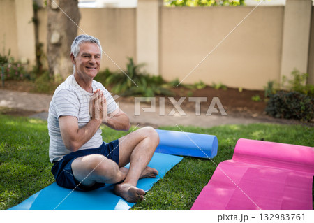 Senior man meditating cross-legged on grass lawn in backyard with blue yoga mat and pink mat Senior man meditating cross-legged on grass lawn in backyard with blue yoga mat and pink mat 132983761
