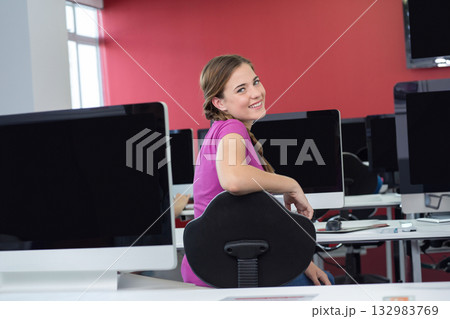 Female student smiling over shoulder while sitting in swivel chair at computer lab with monitors Female student smiling over shoulder while sitting in swivel chair at computer lab with monitors 132983769