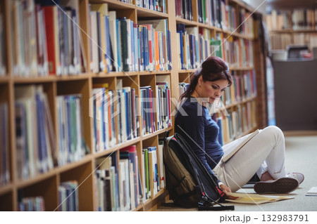 Female student wearing navy top reading book by backpack against bookshelf in library, copy space Female student wearing navy top reading book by backpack against bookshelf in library, copy space 132983791