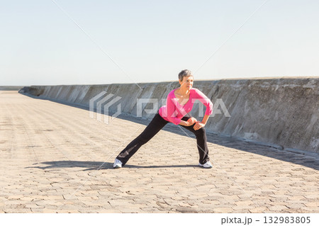 Senior woman in sportswear performing side lunge stretch on seafront walkway by seawall, copy space Senior woman in sportswear performing side lunge stretch on seafront walkway by seawall, copy space 132983805