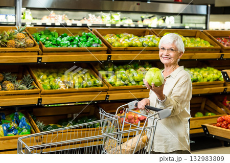 Senior shopper holding green apple while pushing metal cart in grocery produce aisle, copy space Senior shopper holding green apple while pushing metal cart in grocery produce aisle, copy space 132983809