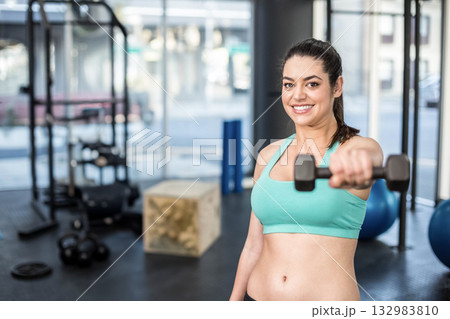 Woman wearing turquoise sportswear doing dumbbell front raise in gym with kettlebells, copy space Woman wearing turquoise sportswear doing dumbbell front raise in gym with kettlebells, copy space 132983810