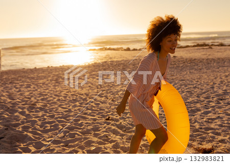 African American woman walking along shoreline at sunset holding yellow inflatable ring, copy space 132983821