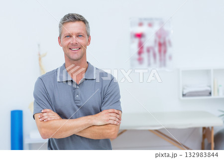 Mature male therapist standing arms crossed in therapy room next to treatment table 132983844