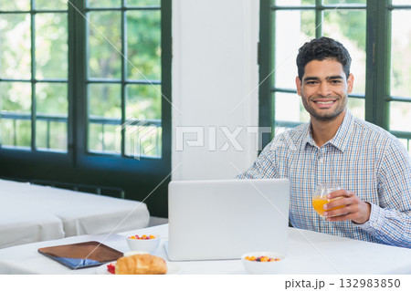 Male office worker holding glass of orange juice while using laptop at breakfast table, copy space 132983850
