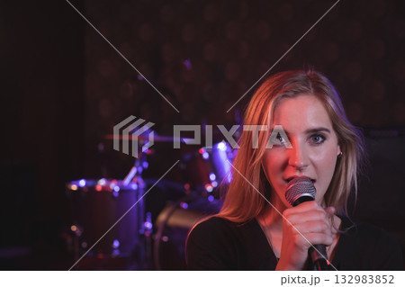 Female singer singing into microphone on stage under purple lights with drum kit, copy space Female singer singing into microphone on stage under purple lights with drum kit, copy space 132983852