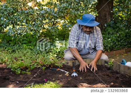 Senior man crouching in home garden spreading soil with trowel in wooden raised bed, copy space Senior man crouching in home garden spreading soil with trowel in wooden raised bed, copy space 132983868