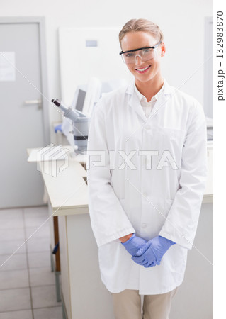 Female scientist wearing goggles, lab coat and gloves at bench in lab using microscope and monitor 132983870