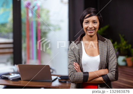 Businesswoman in blazer standing at desk using laptop beside notebooks and pens at glass door 132983876