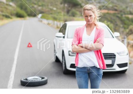 Female driver changing spare tire beside white sedan on hilly road by warning triangle, copy space 132983889