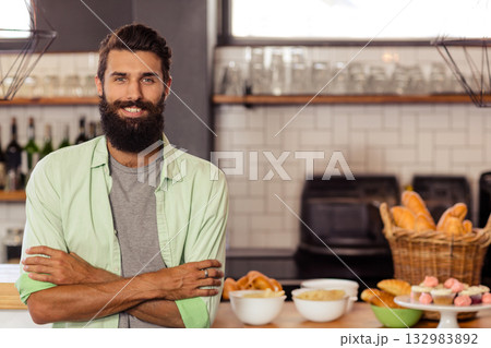 Male baker standing behind counter in bakery showing croissants, baguettes and cupcakes, copy space 132983892