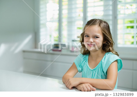 Child girl sitting at white tabletop in bright modern kitchen, smiling under window shutters Child girl sitting at white tabletop in bright modern kitchen, smiling under window shutters 132983899