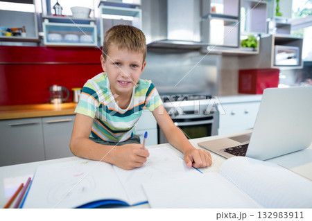 School-age boy writing in notebook with blue pen at kitchen counter with laptop, colored pencils 132983911