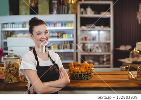 Smiling female barista in black apron serving cookies from jar at counter in bakery, copy space Smiling female barista in black apron serving cookies from jar at counter in bakery, copy space 132983939