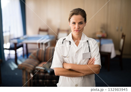 Female nurse standing arms crossed in lounge wearing white scrubs with stethoscope and name badge 132983945
