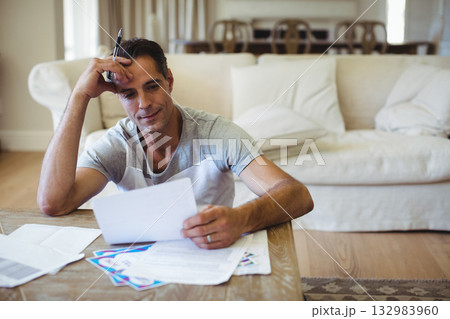 Man sitting on floor in living room using pen reviewing chart papers at low coffee table Man sitting on floor in living room using pen reviewing chart papers at low coffee table 132983960