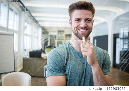 Man wearing green tee holding marker near chin beside flipchart in open-plan office, copy space Man wearing green tee holding marker near chin beside flipchart in open-plan office, copy space 132983985