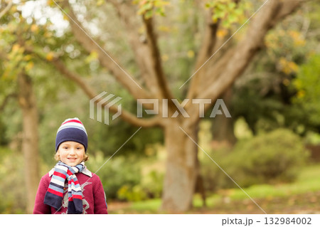 Boy standing, smiling by tree trunk in autumn park, wearing striped knit hat, scarf and sweater Boy standing, smiling by tree trunk in autumn park, wearing striped knit hat, scarf and sweater 132984002
