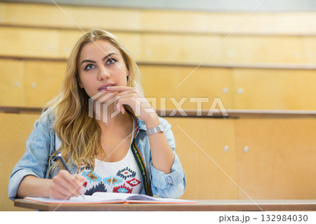 Female student wearing denim jacket writing notes with pen in open notebook on wooden lecture bench 132984030