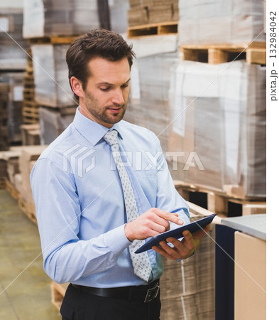 Male supervisor in shirt and tie reviewing inventory on tablet at workstation with boxes on pallets Male supervisor in shirt and tie reviewing inventory on tablet at workstation with boxes on pallets 132984042