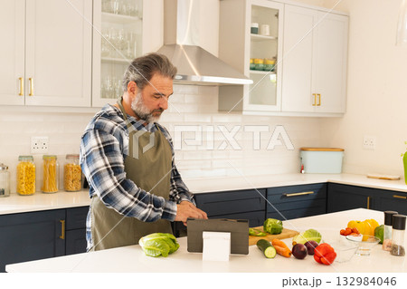 Senior man wearing green apron chopping vegetables on wooden board in kitchen while using tablet Senior man wearing green apron chopping vegetables on wooden board in kitchen while using tablet 132984046
