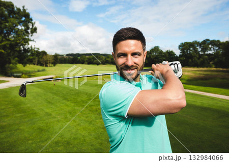 Male golfer wearing turquoise polo and golf glove holding iron club across shoulders on fairway 132984066