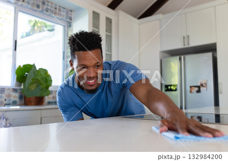 African American man leaning on home kitchen island wiping countertop with cloth under sunny window 132984200