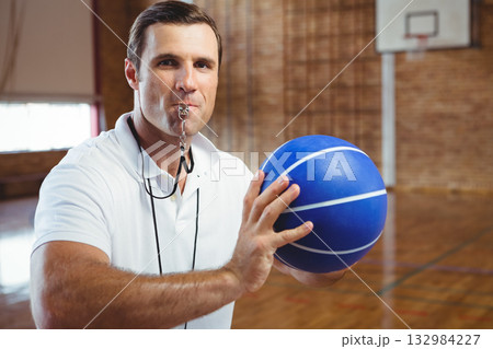 Mature adult male referee holding blue basketball and blowing whistle standing on gymnasium court 132984227