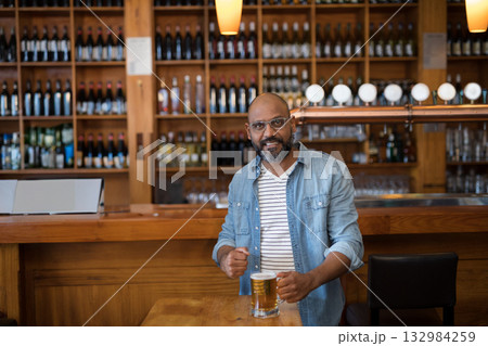 Indian man standing at bar counter holding glass beer mug under warm pendant lights 132984259