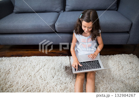 Girl child sitting on white rug in front of dark grey sofa working on silver laptop Girl child sitting on white rug in front of dark grey sofa working on silver laptop 132984267